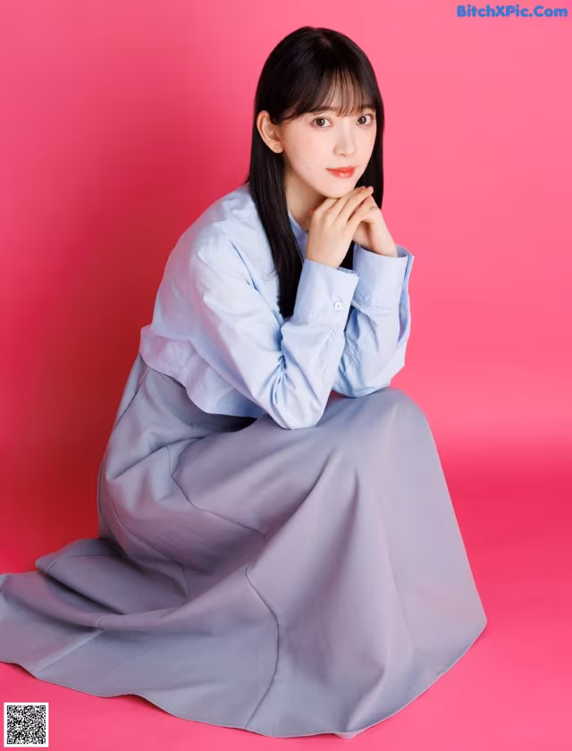 A woman in a blue shirt and gray skirt sitting on a pink background.